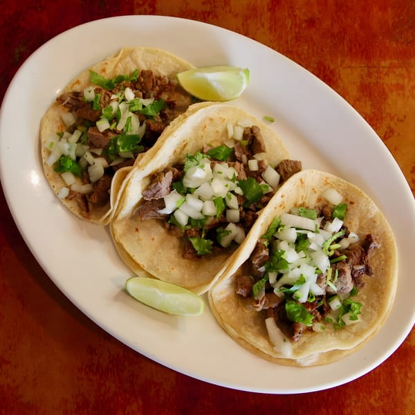 Three street tacos with meat, diced onions, cilantro, and lime wedges on a white plate.