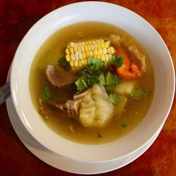 Hearty soup in a white bowl with corn, beef, cabbage, carrots, and cilantro on a red table.